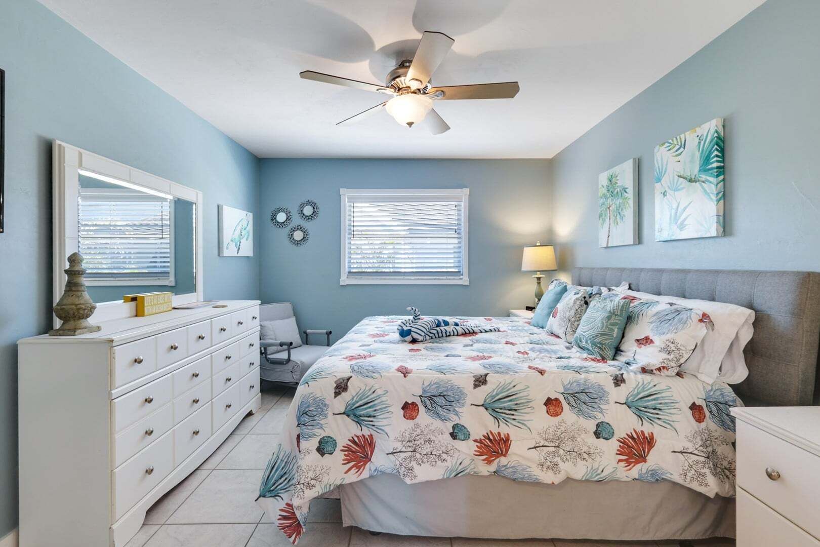 A bedroom with a bed, dresser, chair, ceiling fan, and wall art.