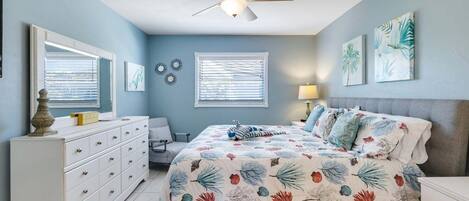 A bedroom with a bed, dresser, chair, ceiling fan, and wall art.