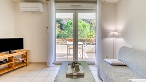 Una sala de estar con televisor de pantalla plana, mueble de madera para entretenimiento, un sofá, una mesa de centro y un ventanal corredizo que da a un balcón exterior.