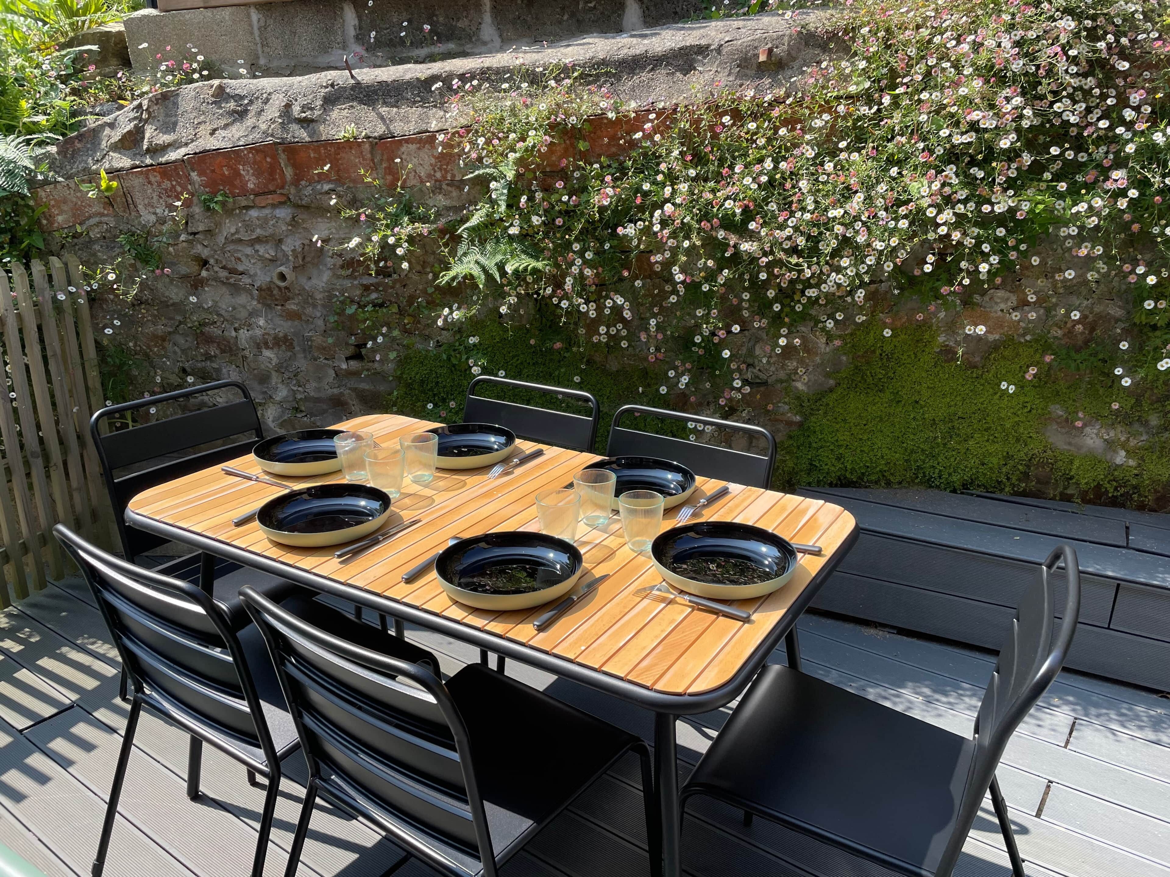 An outdoor dining area with a wooden table and black chairs, set against a stone wall with flowering plants.