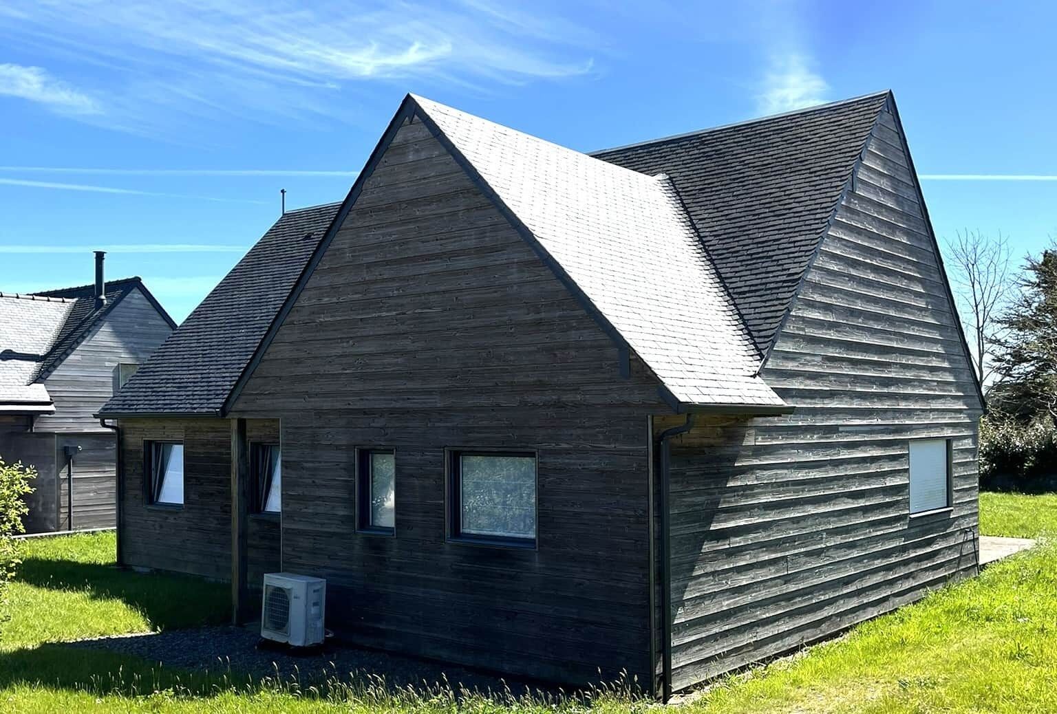 A modern wooden house with a gabled roof, a small attached garage, and a visible air conditioning unit.