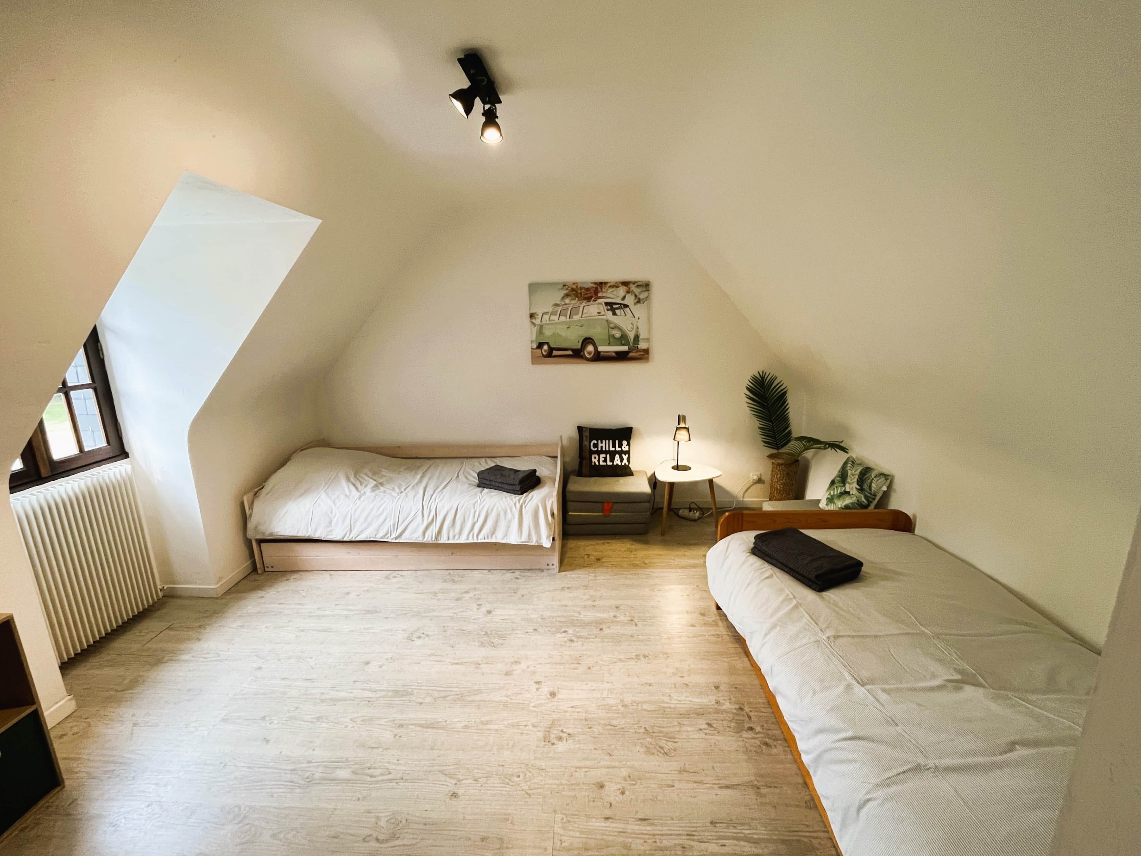 Une chambre avec deux lits, un radiateur, un luminaire au plafond, une petite table avec une lampe, une plante et un tableau accroché au mur.