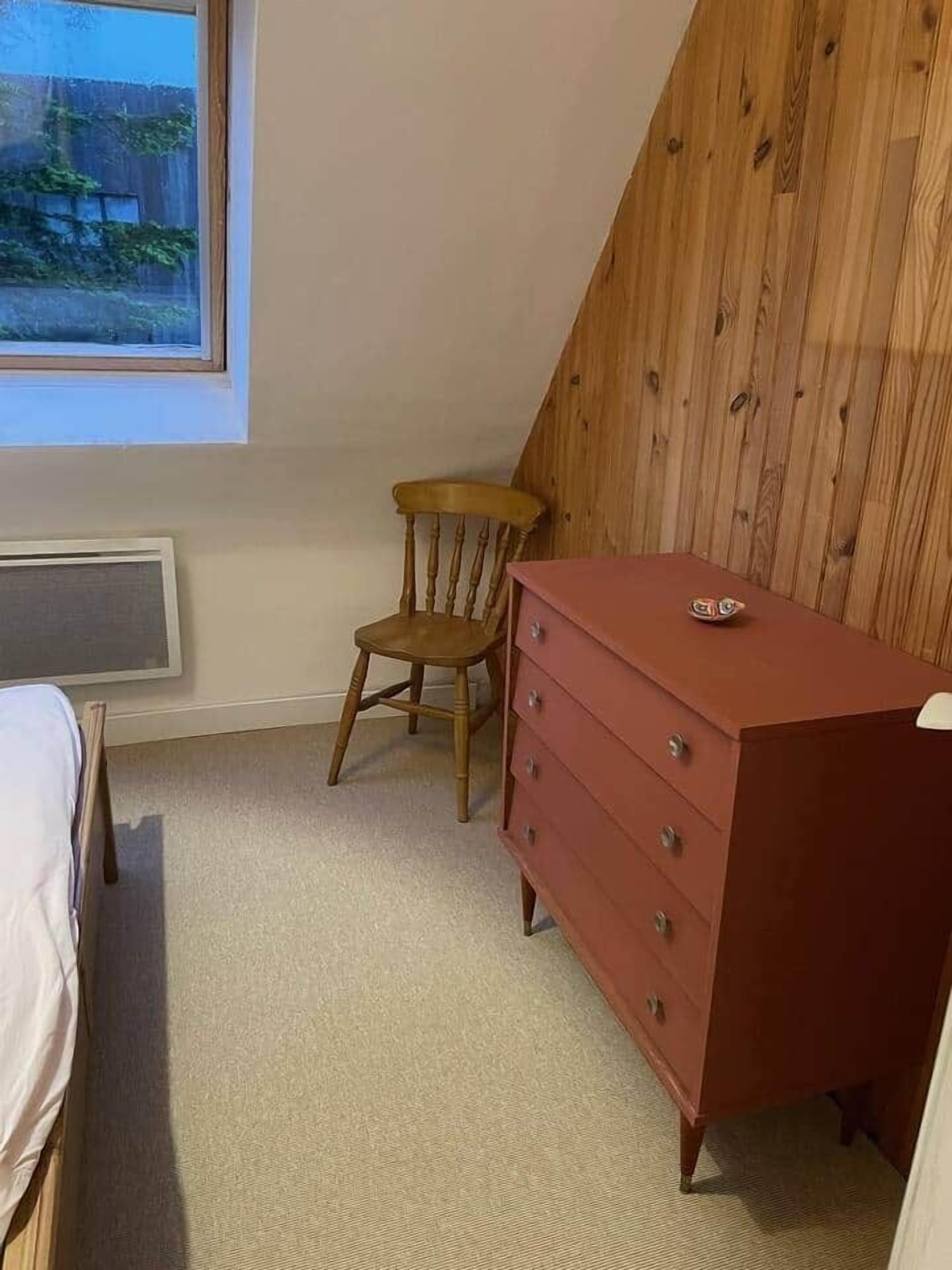 A bedroom with a wooden sloped ceiling, a wooden chair, a red dresser, and a window with a view of a fence and greenery.