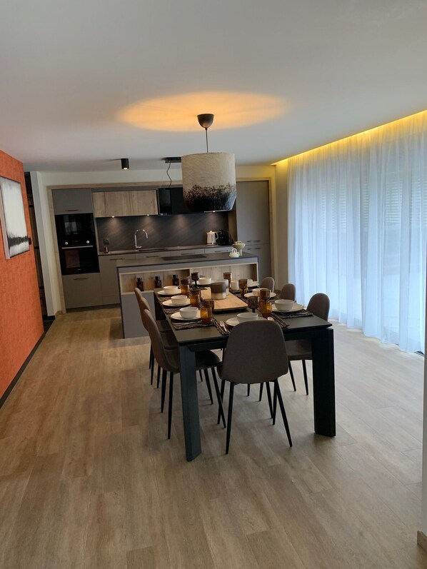 A modern kitchen with a central island, a dining area with a table set for four, and a large window with sheer curtains.