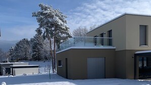 Una casa moderna con un patio cubierto de nieve, un garaje y un árbol con ramas nevadas.