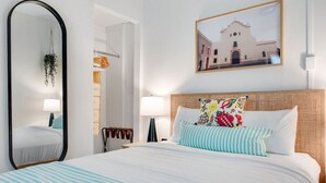 Una habitación de hotel con una cama grande, un espejo, un cuadro de un edificio y una planta colgante.