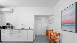 A modern kitchen with white cabinets, a microwave, and a sink. There is a dining area with orange chairs and a small table. A painting of a sunset is on the wall.
