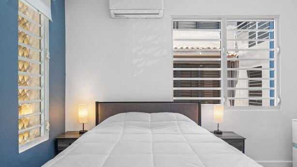 A bedroom with a large bed, two bedside tables with lamps, a window with blinds, and an air conditioning unit on the wall.