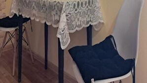 Un comedor con una mesa cubierta por un mantel de encaje, dos sillas y un cartel en la pared que anuncia un club de café y un club de vino y queso.