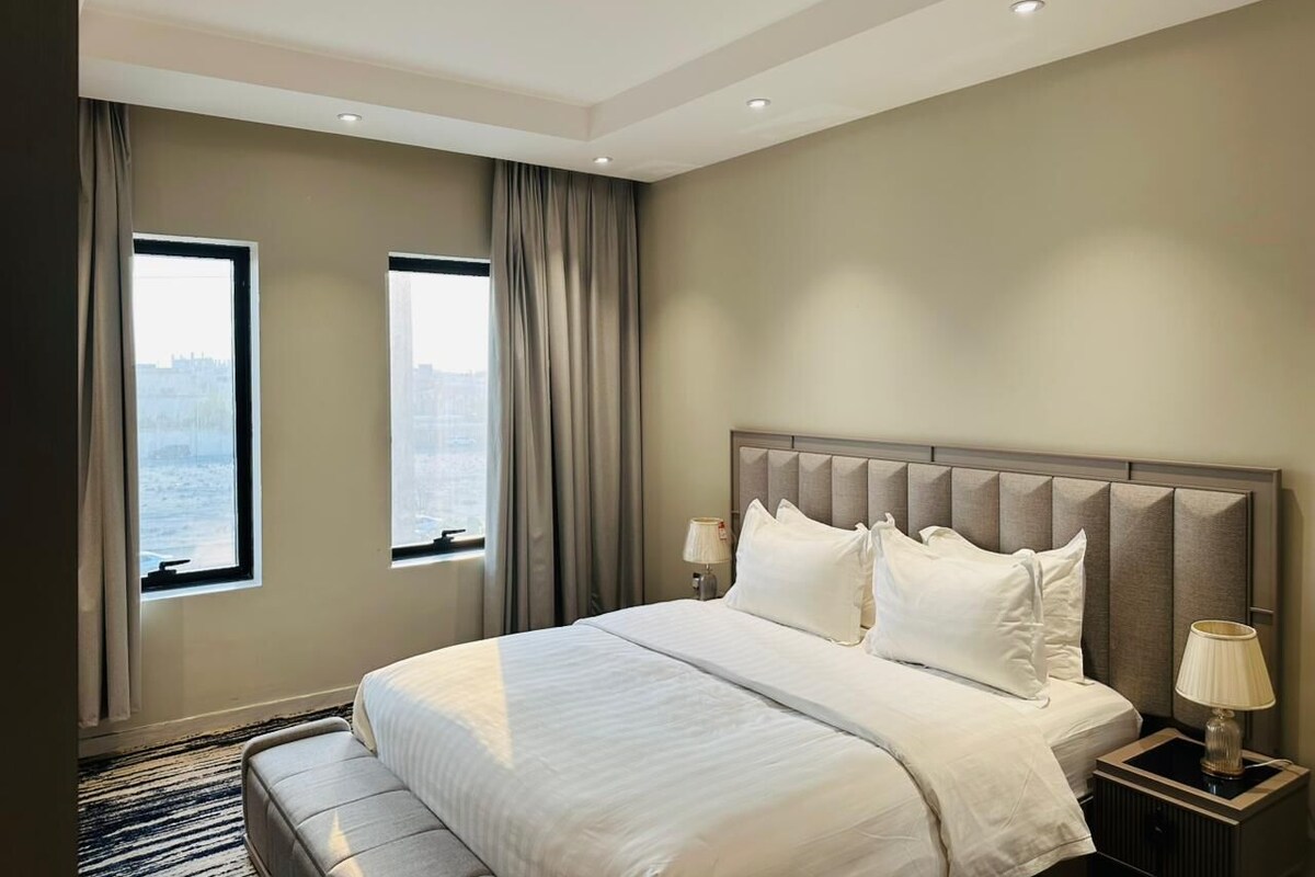 A hotel room with a large bed, two windows with curtains, a headboard, and bedside lamps.