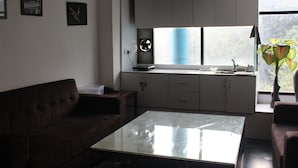 A modern living room with a glass coffee table, a dark brown sofa, and a kitchen area with white cabinets and a window. - Flourish Inn Saket opposite Max Hospital (New Delhi)