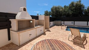 Un'area cucina esterna con barbecue, lavello e pensili, un tavolo rotondo in legno e una sedia a bordo piscina.