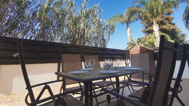 Patio esterno con tavolo apparecchiato per consumare pasti, circondato da sedie e affacciato su un giardino con erba alta e palme.