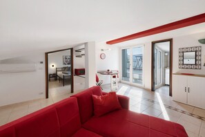 A modern living room with a red sofa, a dining area with a table and chairs, a kitchen with cabinets, and a large window providing natural light.