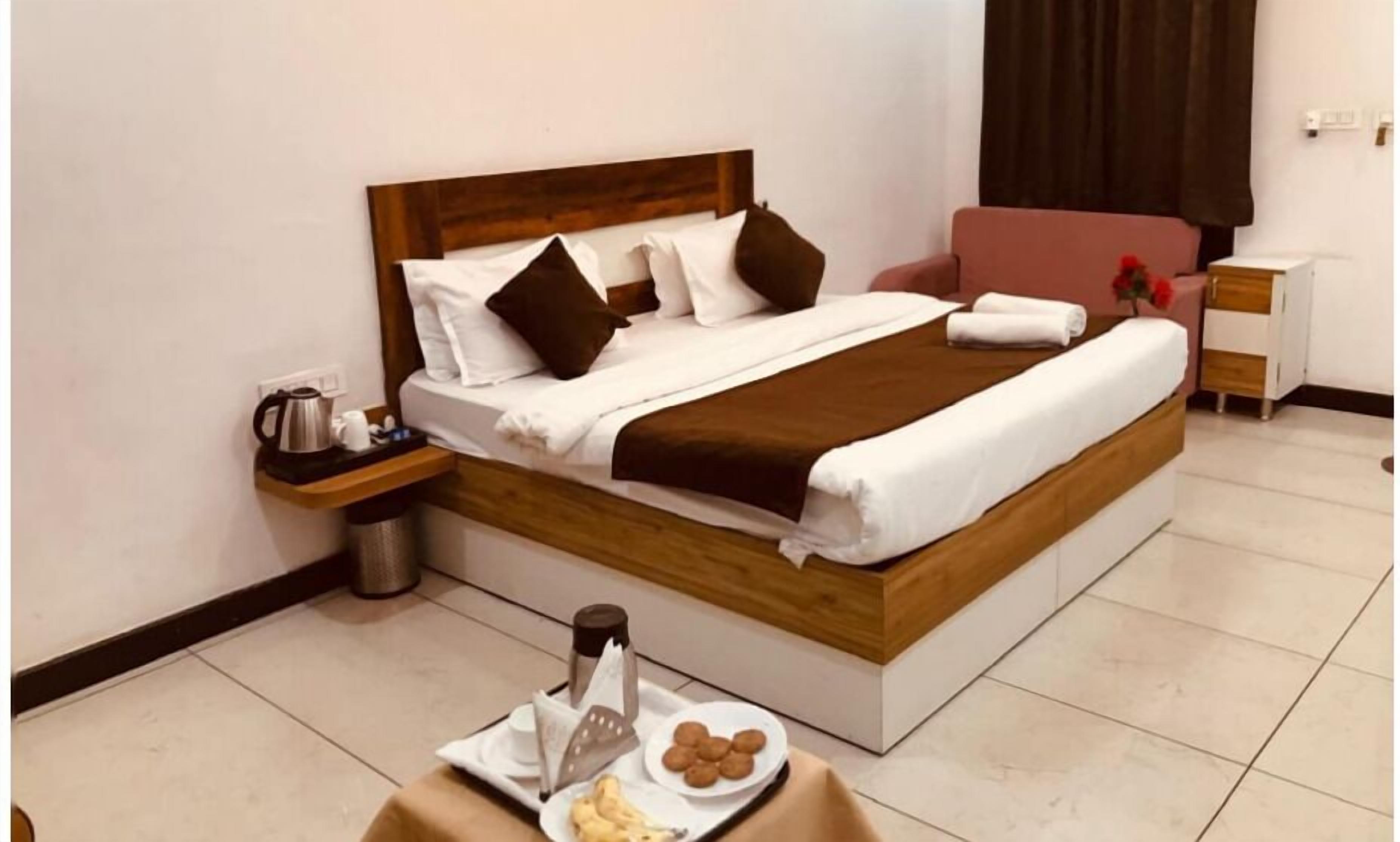 Una cama bien hecha con cabecera de madera, una mesita de noche con hervidor y tazas, y una bandeja con comida y una botella.