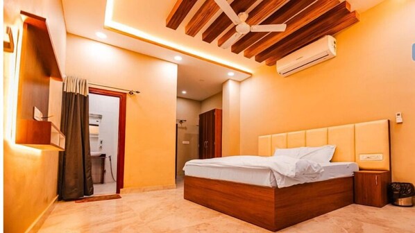 A modern bedroom with a wooden headboard, a bed with white linens, a built-in air conditioning unit, and a shower area. - Umraogarh A Unit of Raghav Vilas (Jodhpur)