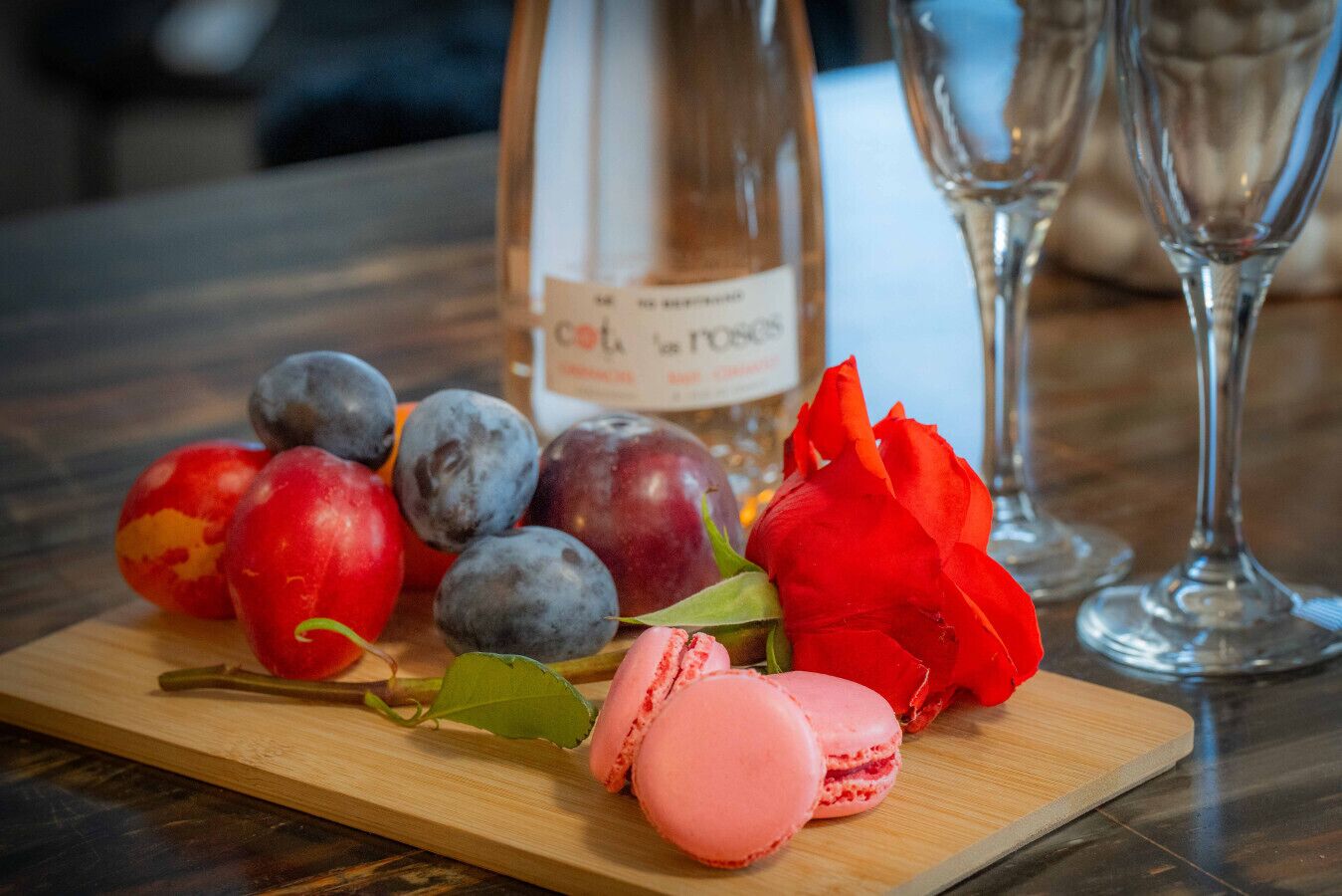A wooden cutting board with fruits, macarons, and a bottle of wine on a table.