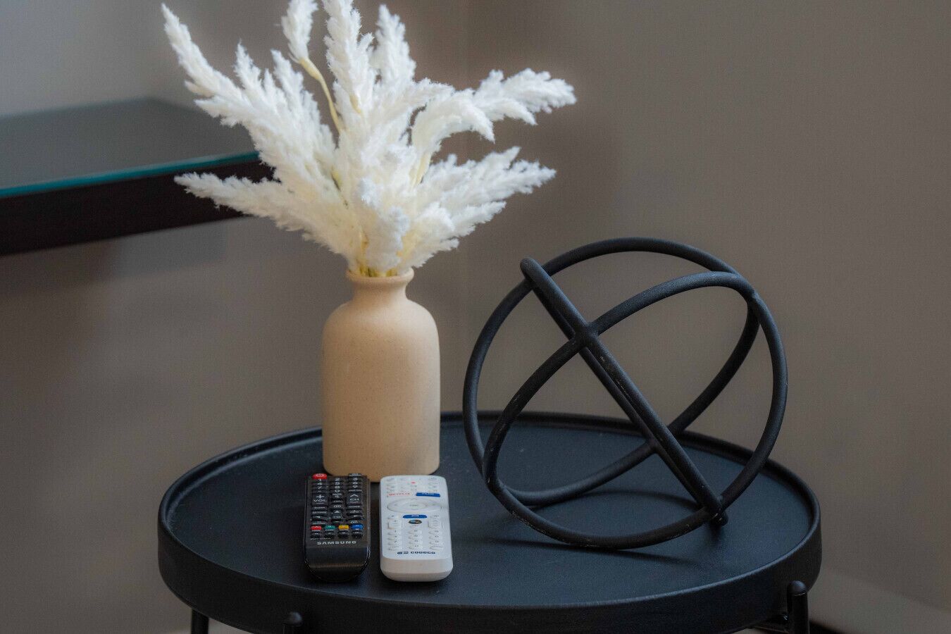 A small round table with a vase of white flowers, two remote controls, and a black geometric object.