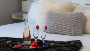 A bed with a white pillow and a fluffy grey pillow, a tray with a bottle, glasses, strawberries, and fruits.