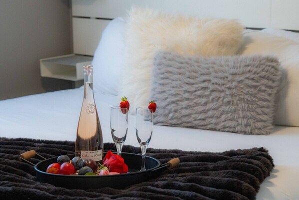 A bed with a white pillow and a fluffy grey pillow, a tray with a bottle, glasses, strawberries, and fruits.