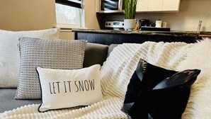 A cozy living room with a grey sofa, a white knitted throw, and a pillow with the text "LET IT SNOW". - 8 KING Bed, Comfy Sofa, Close To Attractions (Niagara Falls)