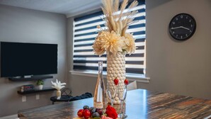 A wooden dining table with a decorative vase, fruit, and a bottle. A clock and a television are visible in the background. - 8 KING Bed, Comfy Sofa, Close To Attractions (Niagara Falls)