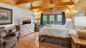 Ein Schlafzimmer einer Holzhütte mit Bett, Kommode, Fernseher und einem großen Fenster mit Blick auf Grün.
