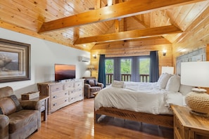 A wooden cabin bedroom with a bed, dresser, TV, and a large window with a view of greenery. - Lodge with Fire Pit, Indoor and Outdoor Fireplace, Hot Tub, Games, King Beds (Sevierville)