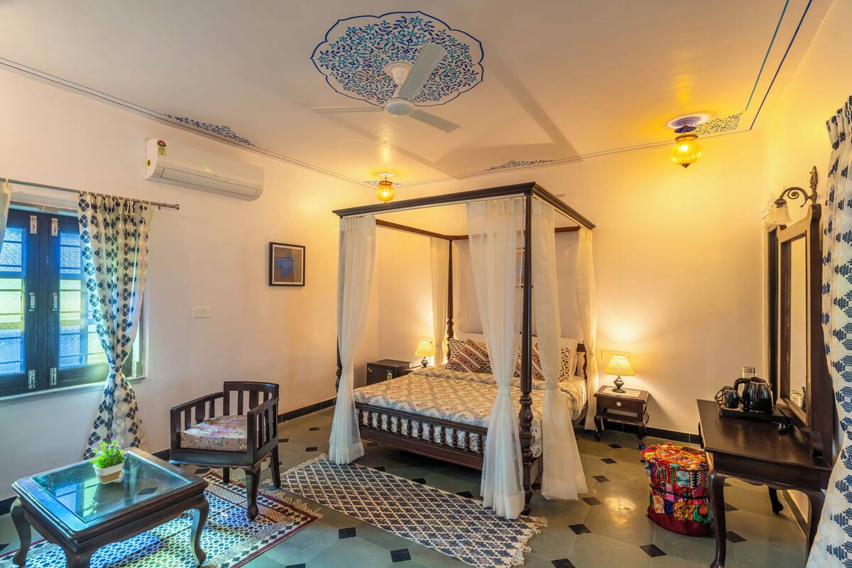A four-poster bed with a canopy, a wooden nightstand with a lamp, a small table with a glass top, and a chair with a floral cushion.