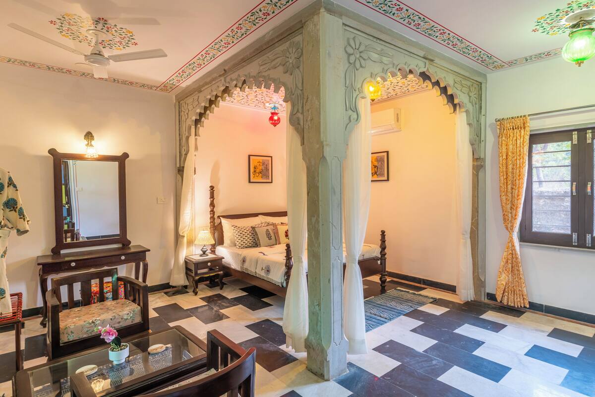 A traditional room with a bed, a sitting area with a table and chairs, a mirror, and a decorative ceiling fan.