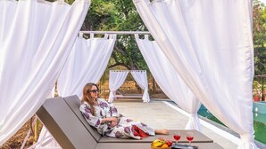 Una persona descansando en una tumbona con una mesa cerca, una cesta de fruta y dos copas de vino tinto.