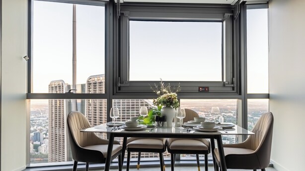 Een eetruimte met een tafel voor vier personen, een vaas met bloemen en een stadszicht door grote ramen.