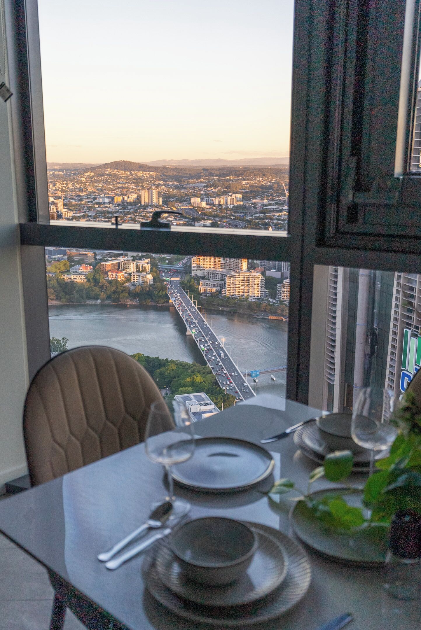 A dining table set with plates, cutlery, and a glass of wine, with a view of a cityscape and river through a window.