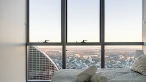 Een slaapkamer met een groot raam, een bed met een opgerolde handdoek en uitzicht op een stadsbeeld.