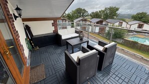 Un espace extérieur aménagé, abrité et doté de mobilier en osier, d’une balustrade en verre et offrant une vue sur des maisons et une piscine.