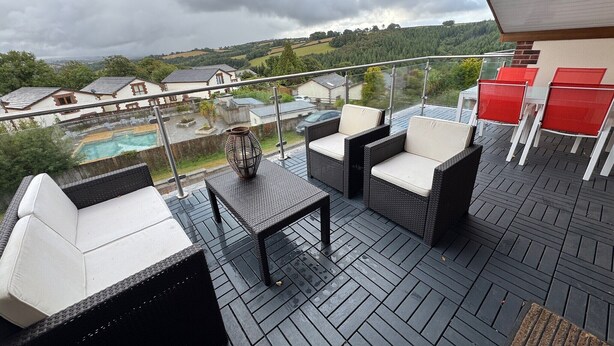 Un balcon doté de meubles en osier, d’une rambarde en verre et offrant une vue sur une piscine et des maisons.
