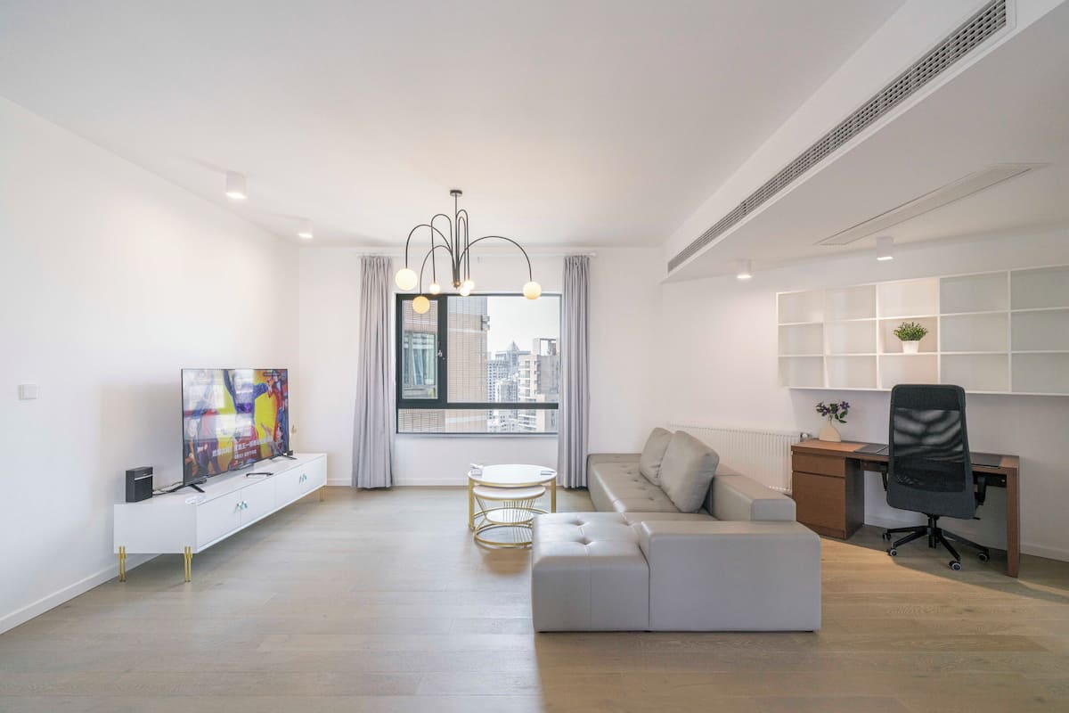 A modern living room with a white sofa, a TV, a dining table, and a city view through large windows.