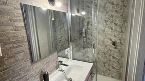 Une salle de bain moderne avec un mur à l’aspect pierre, un grand miroir, un lavabo blanc et une douche à parois de verre.