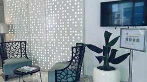 A modern waiting area with a sofa, chairs, a table, a potted plant, a TV, and a framed informational sign. - The Mayberry A Bergamont Hotel Tiruppur (Tirupur)