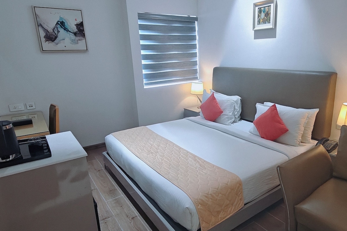 Ein Hotelzimmer mit Bett, Nachttischlampen, einem Sessel, einem Schreibtisch mit Kaffeemaschine und einem Fenster mit Jalousien.