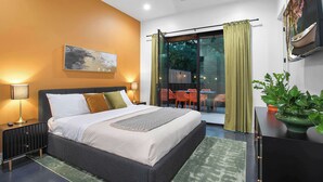 Un dormitorio moderno con una cama grande, una mesita de noche con lámpara, una planta verde y vistas a un área de estar exterior.