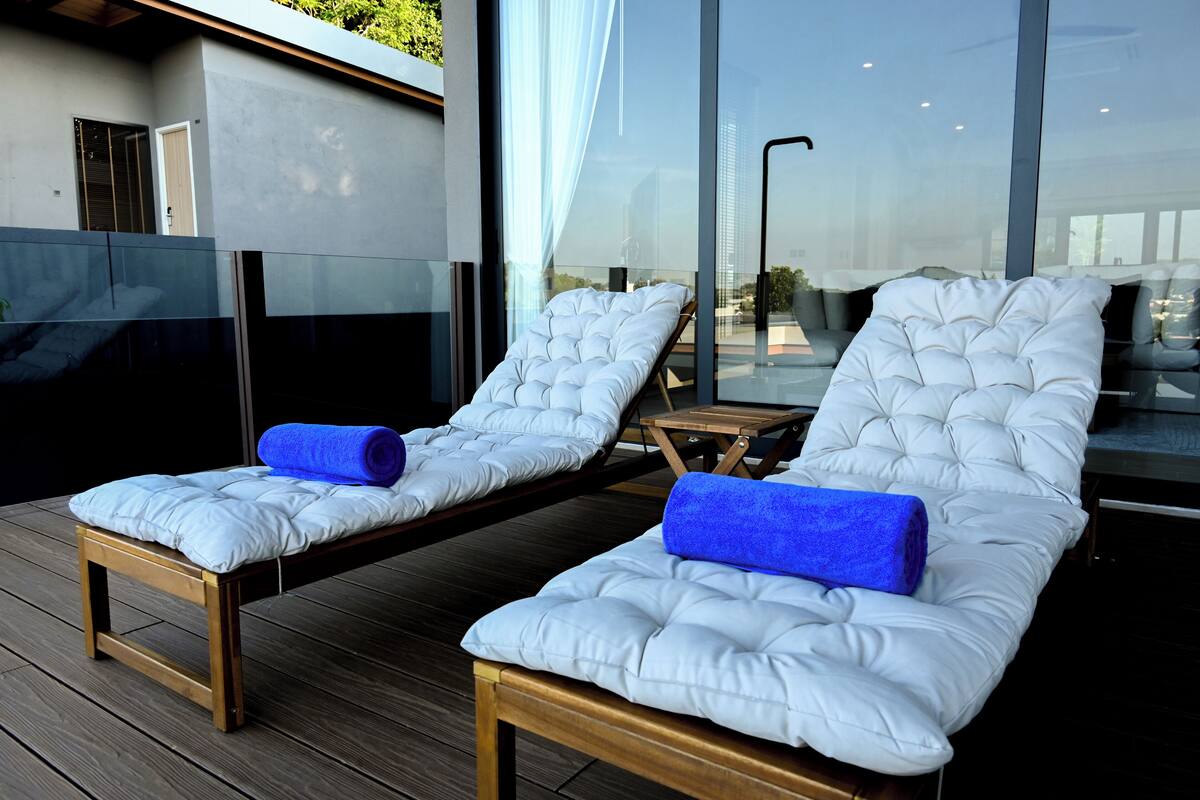 Dos sillones blancos con cojines y toallas azules sobre una terraza de madera, con vista a un edificio moderno con amplias ventanas.