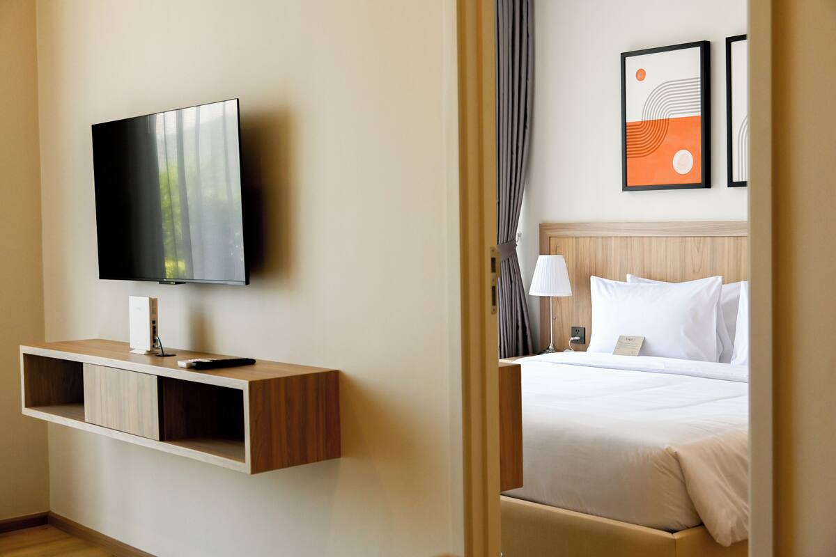 Habitación de hotel con una televisión de pantalla plana montada en una consola de madera, una cama con ropa blanca, una mesita de noche con lámpara y dos cuadros en la pared.
