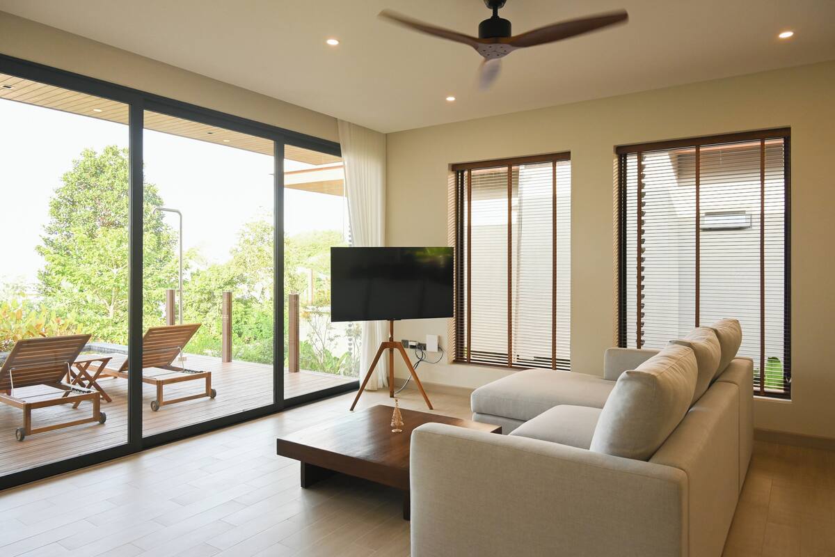 Un salón moderno con un sofá, un televisor sobre un mueble bajo y un balcón con sillas.