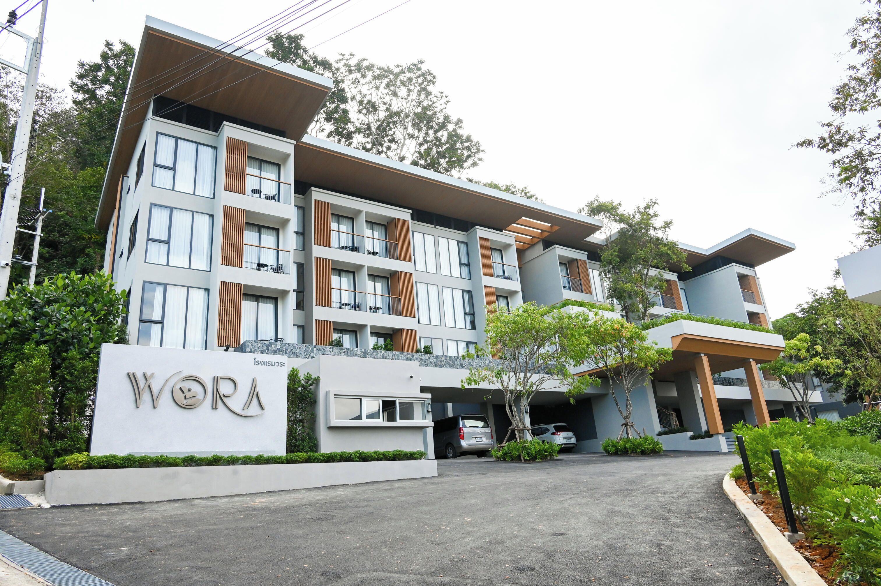 Un hotel moderno de varios pisos con balcones de listones de madera, un área de estacionamiento y un letrero con el nombre 'WORA'.
