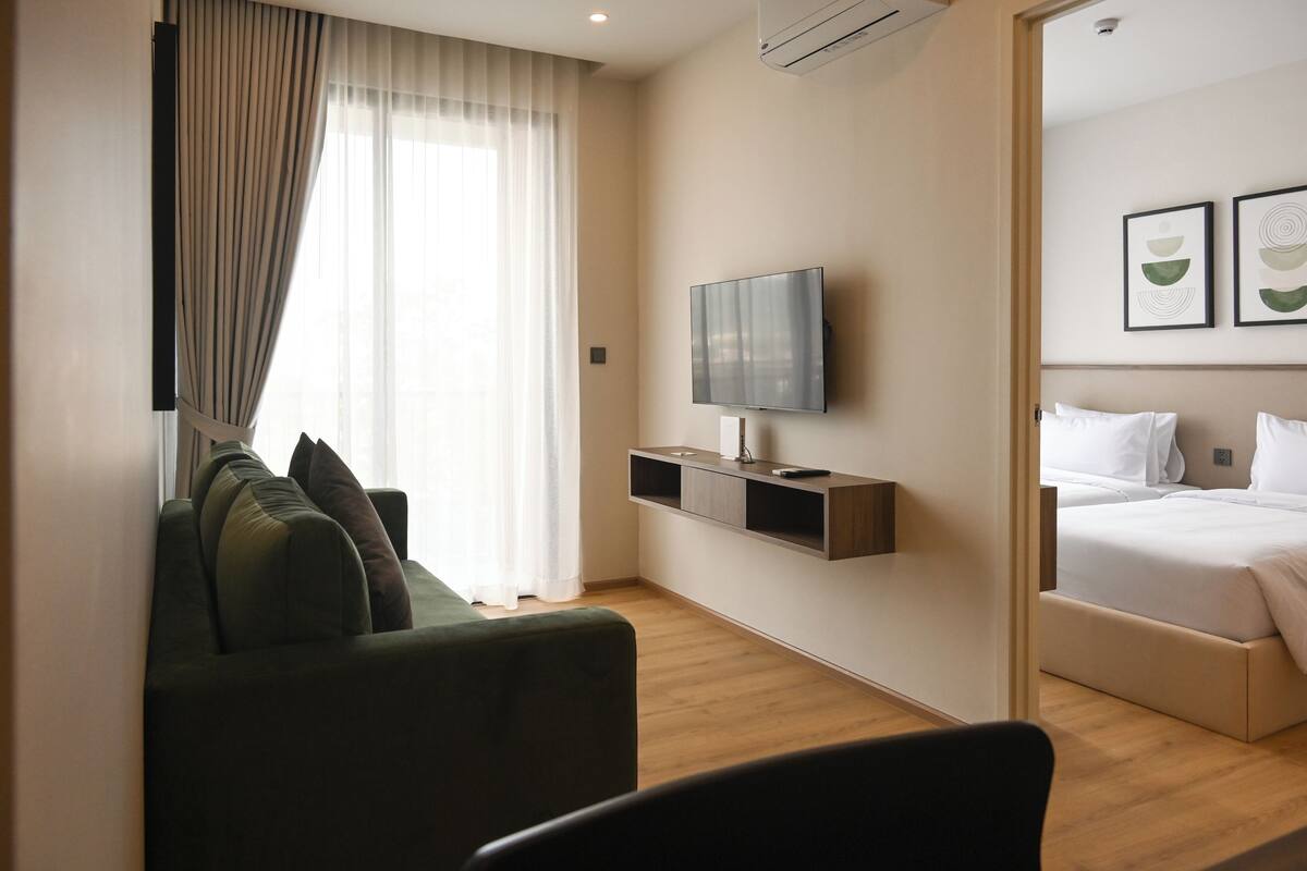 Una habitación de hotel moderna con sofá, televisor montado en la pared, cama y ventana con cortinas.