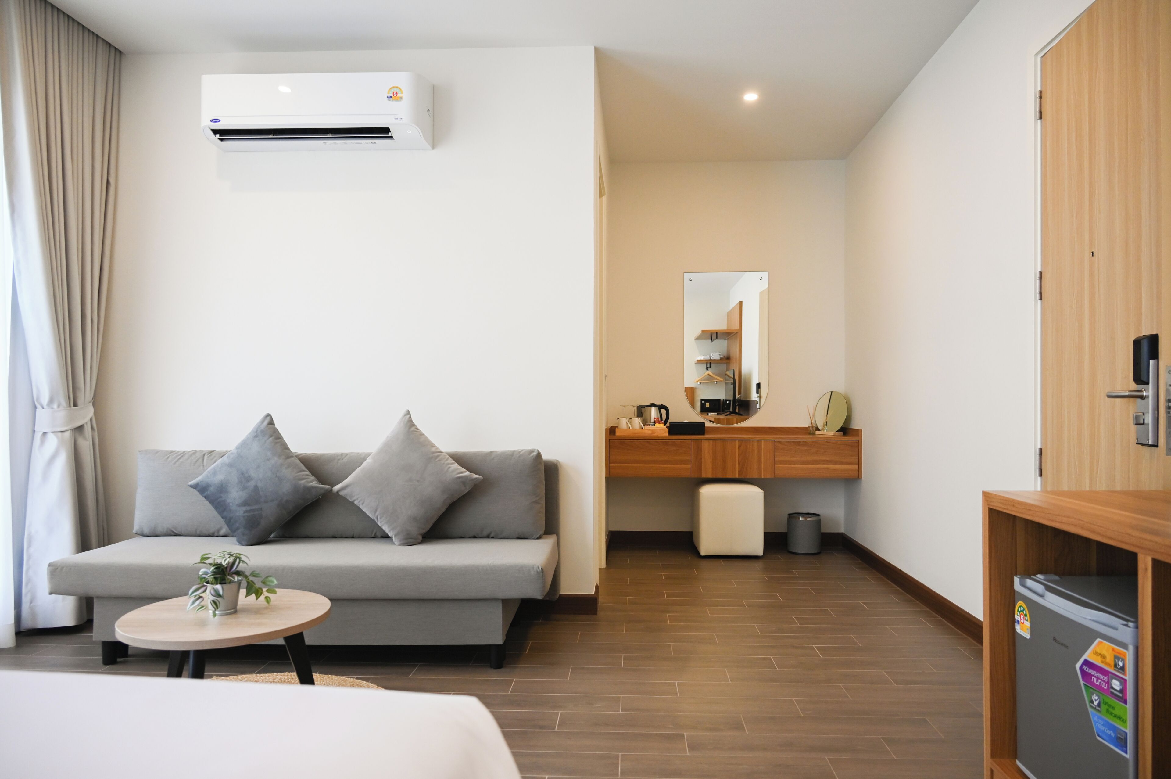 Een moderne hotelkamer met een grijze bank, een klein rond tafeltje met een plant, een houten kast, een spiegel en een inbouwkoelkast.