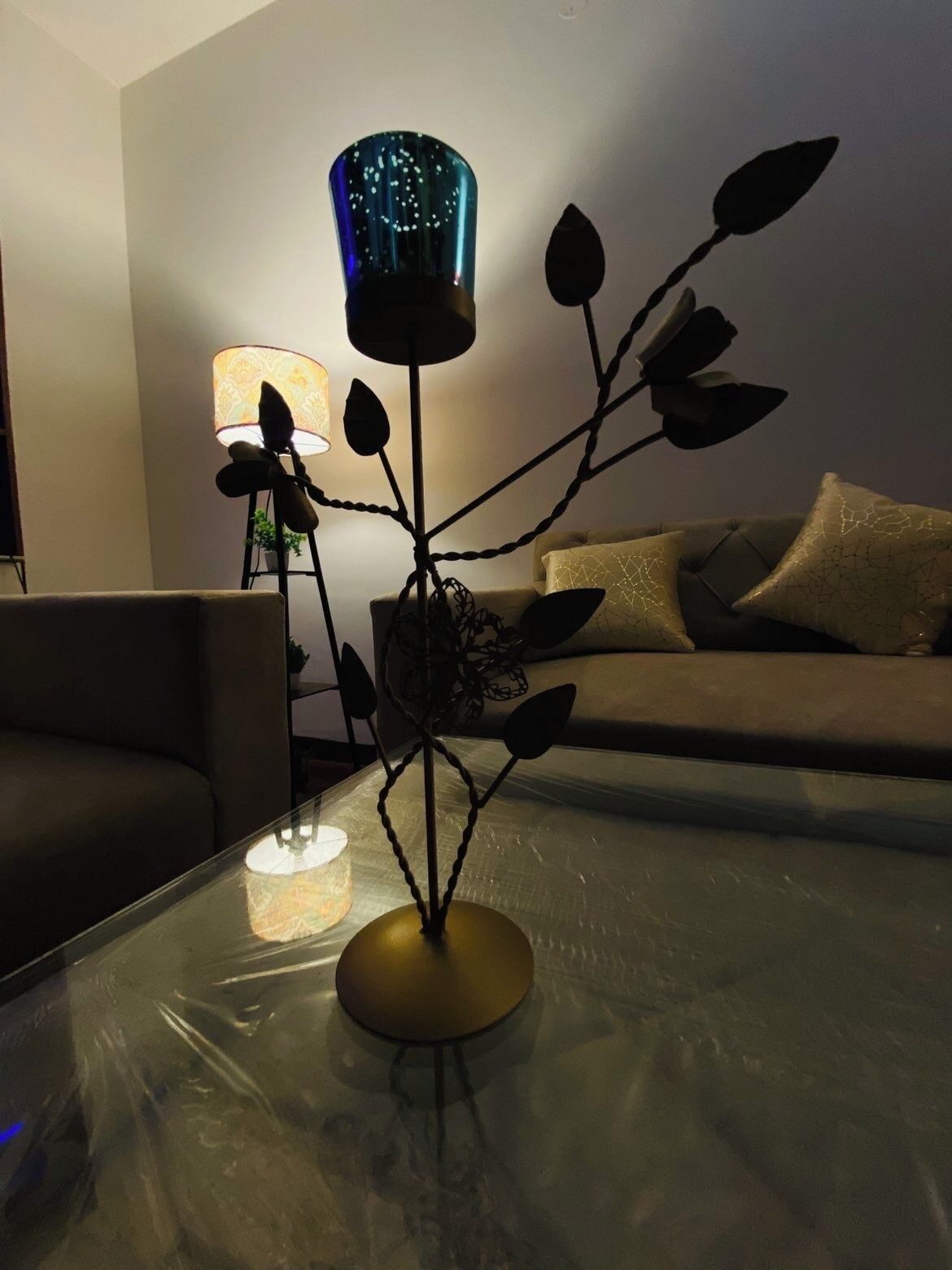 Un salon avec une table basse en verre, une plante décorative en métal, une lampe bleue et un canapé avec des coussins.