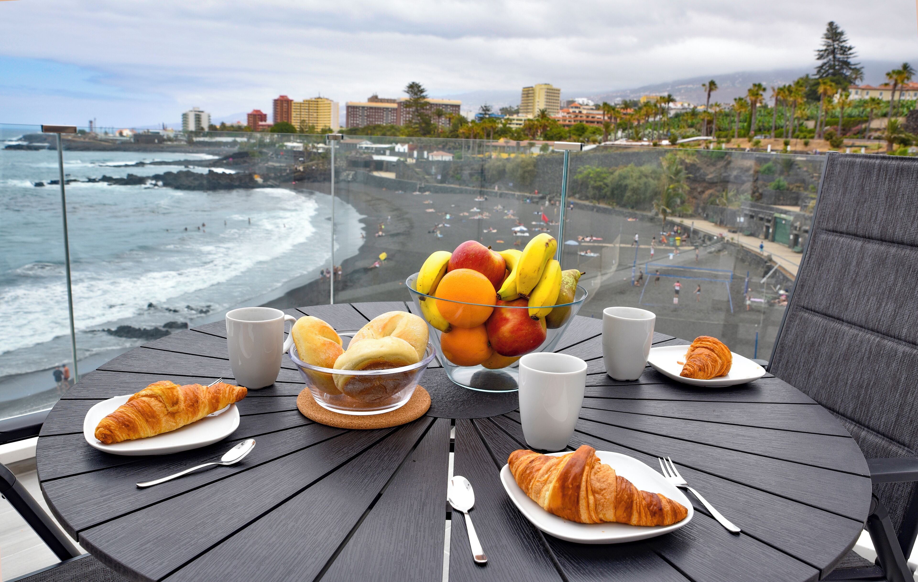 Una mesa con croissants, fruta y café, con vista a una playa y edificios al fondo.