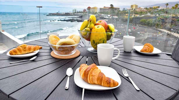 A table with croissants, a fruit bowl, and a cup, overlooking a coastal view.